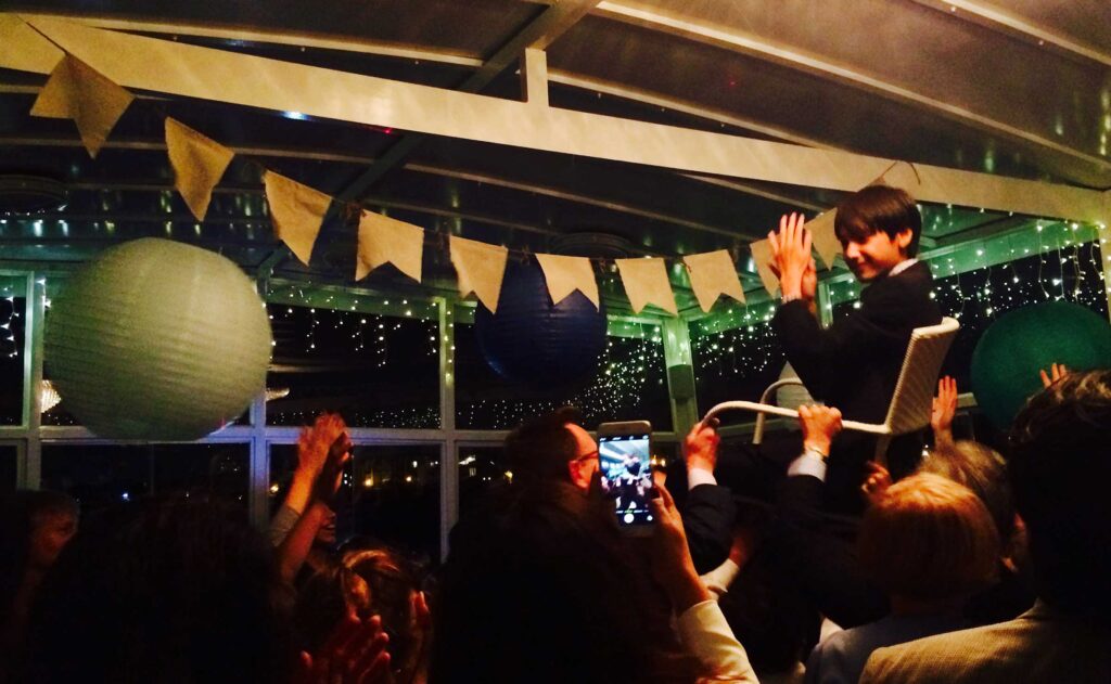 Bar/Bat Mitzvah Party celebration. Young boy hoisted in a chair celebrating. Banner in background, on a yacht.